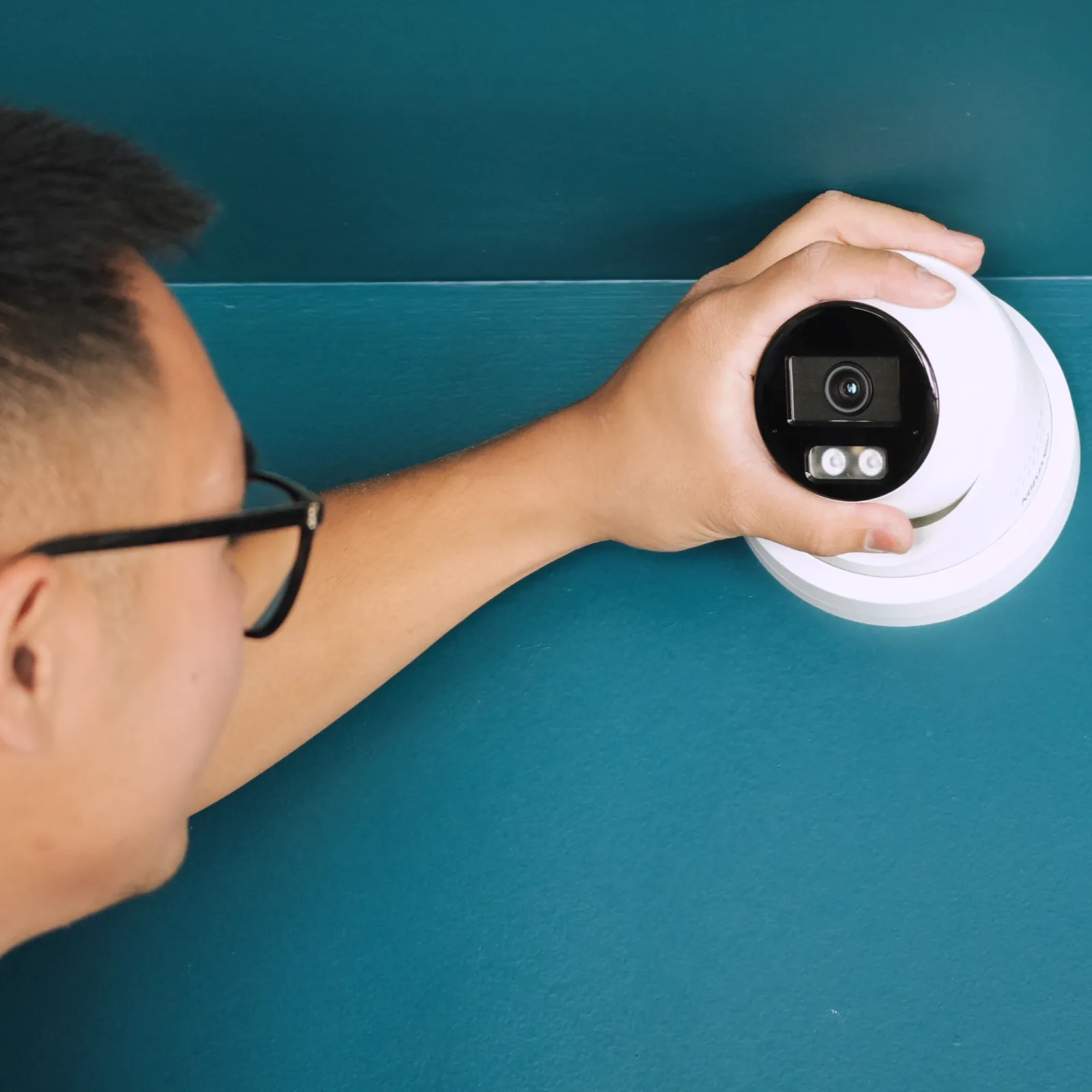 Technicien SERELYS SÉCURITÉ en Vendée installant une caméra dôme IP de vidéosurveillance blanche avec éclairage LED intégré sur un mur teal