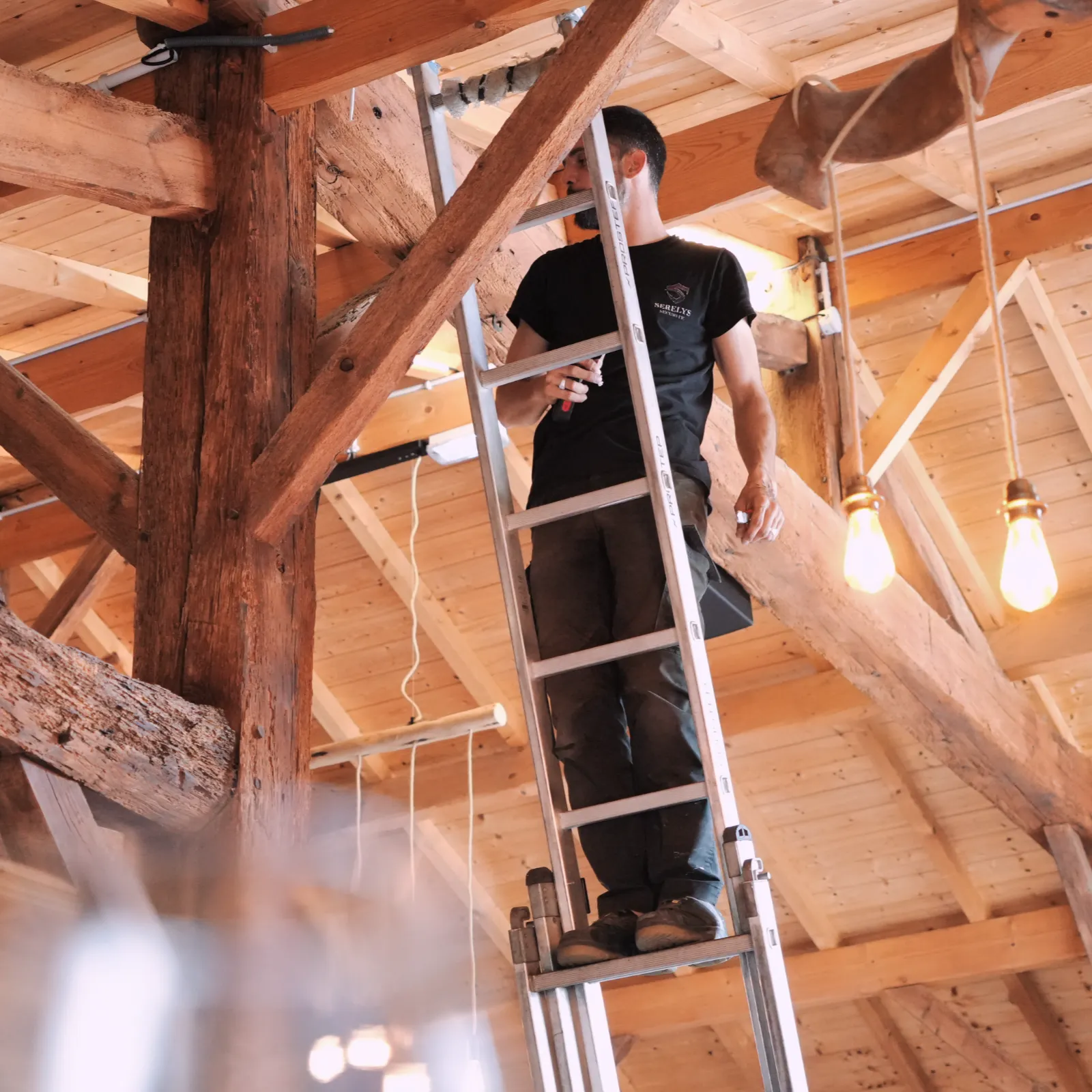 Technicien SERELYS SÉCURITÉ en Vendée installant un système de vidéosurveillance dans une structure en bois, grimpant sur une échelle pour positionner équipements de sécurité