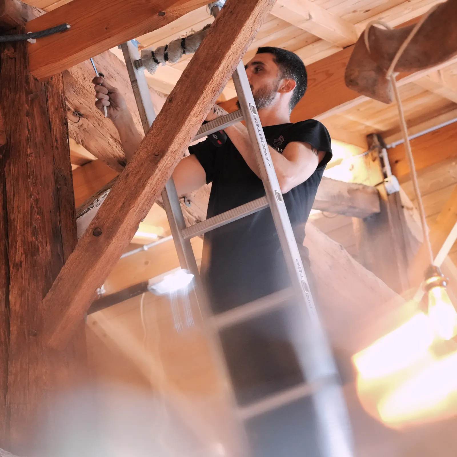 Technicien SERELYS SÉCURITÉ en Vendée effectuant l'installation de caméras de vidéosurveillance dans les combles d'un bâtiment, utilisant une échelle pour accéder aux structures en bois