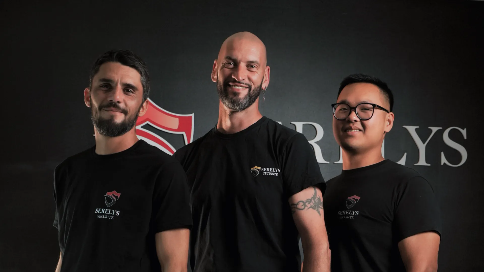Trois professionnels de SERELYS SÉCURITÉ en Vendée portant des t-shirts noirs avec le logo de l'entreprise, posant devant un fond gris avec le nom SERELYS visible