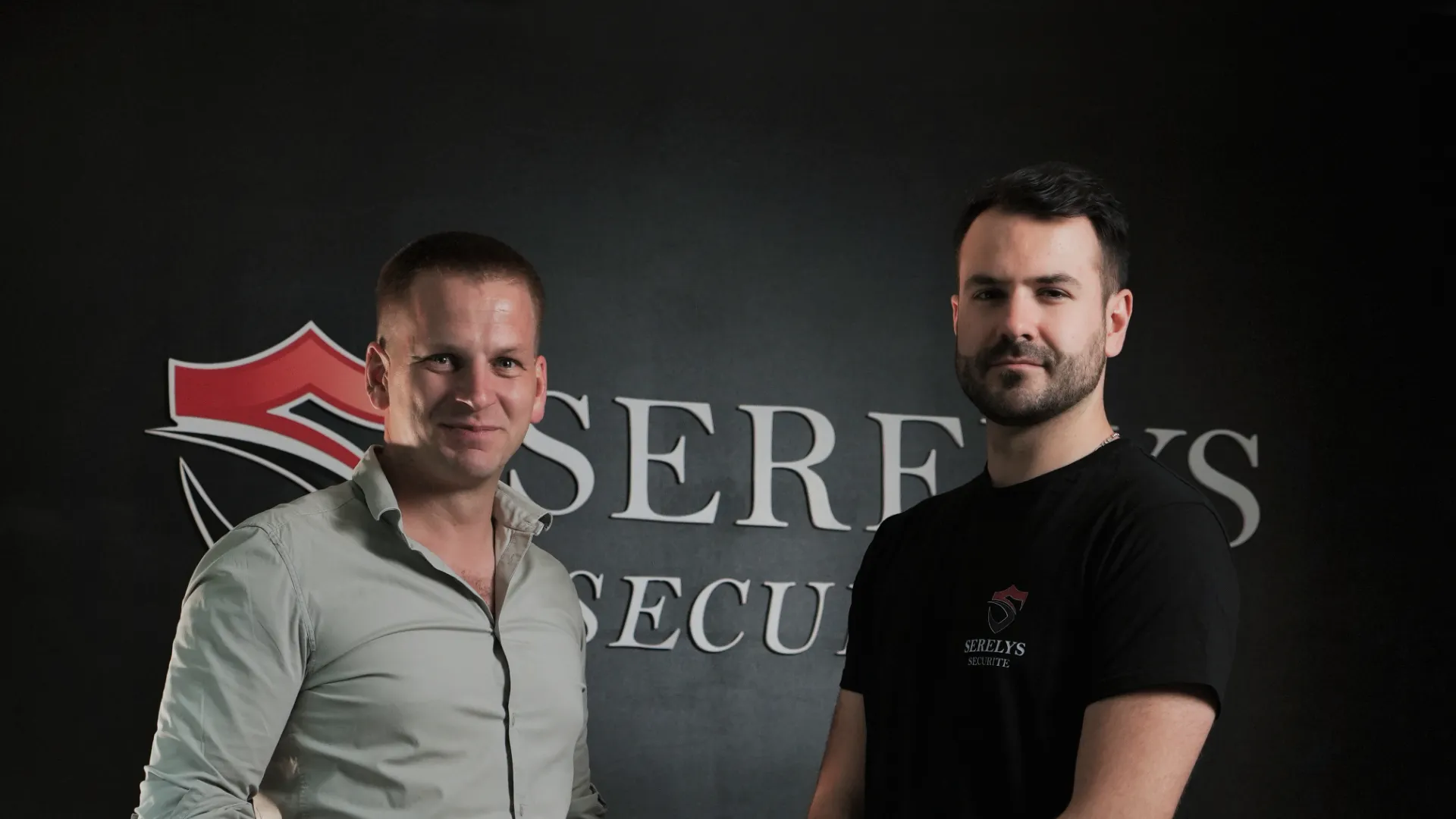 Deux professionnels de SERELYS SÉCURITÉ en Vendée posent devant le logo de l'entreprise, représentant l'équipe expert en vidéosurveillance et solutions de sécurité