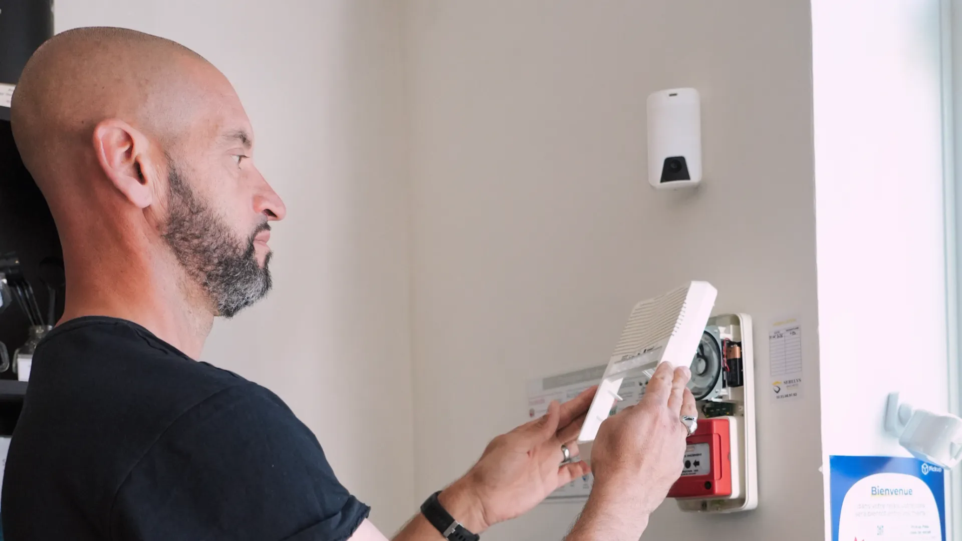 Technicien SERELYS SÉCURITÉ en Vendée installant une détecteur de mouvement PIR blanc sur mur blanc, avec panneau électrique et alarme incendie rouge visibles