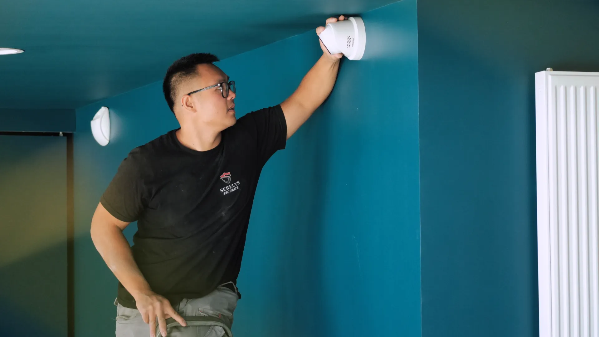 Technicien SERELYS SÉCURITÉ en Vendée installant un détecteur de fumée blanc au plafond d'une pièce aux murs bleu turquoise