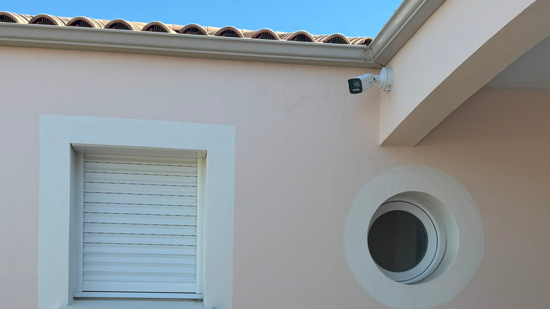 Installation de vidéosurveillance SERELYS SÉCURITÉ en Vendée : caméra de surveillance fixée à l'angle d'une façade blanche avec store et bouche d'aération