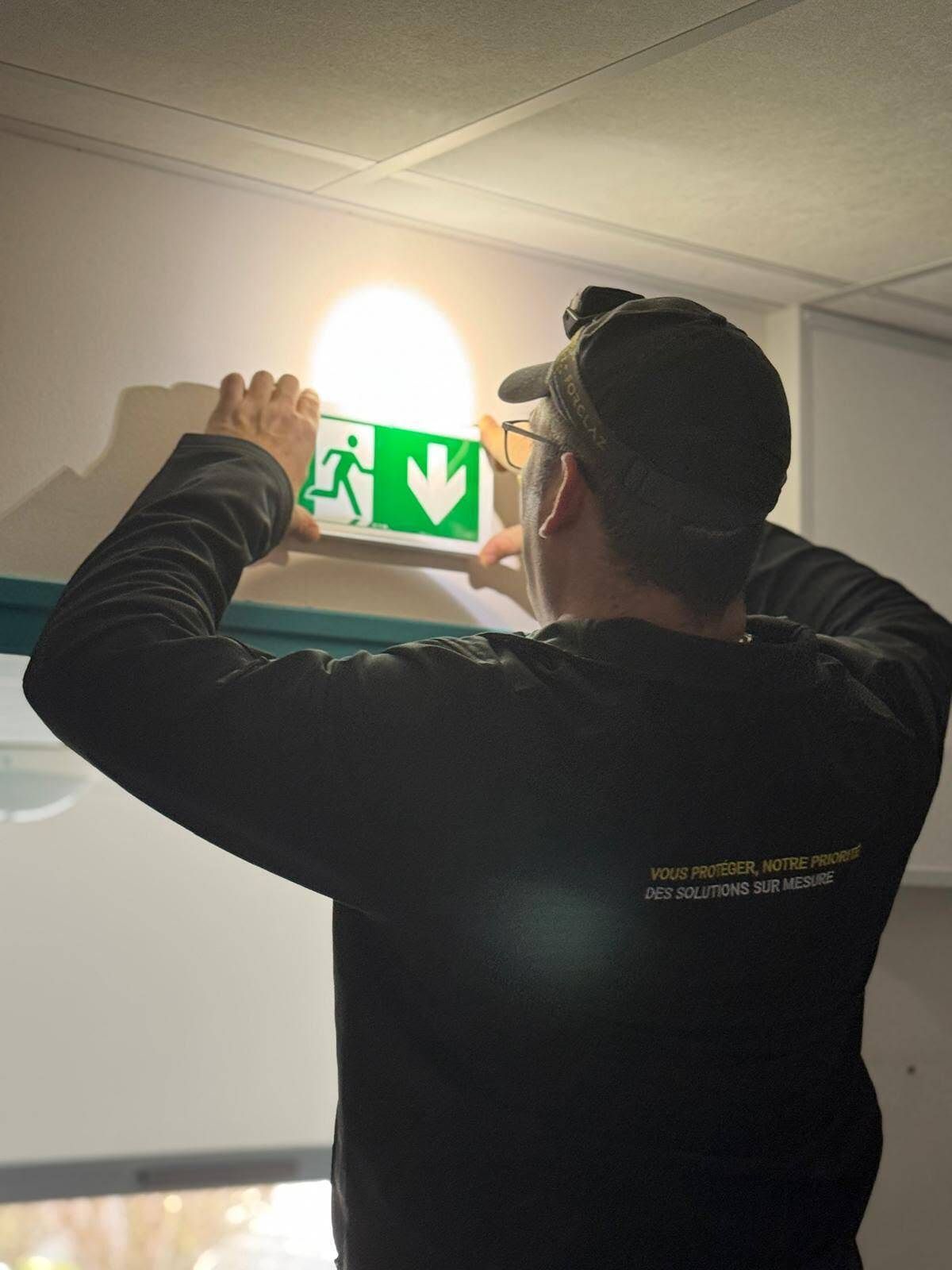 Technicien SERELYS SÉCURITÉ en Vendée installant une signalétique d'évacuation lumineuse verte dans un bâtiment commercial, démontrant son expertise en solutions de sécurité